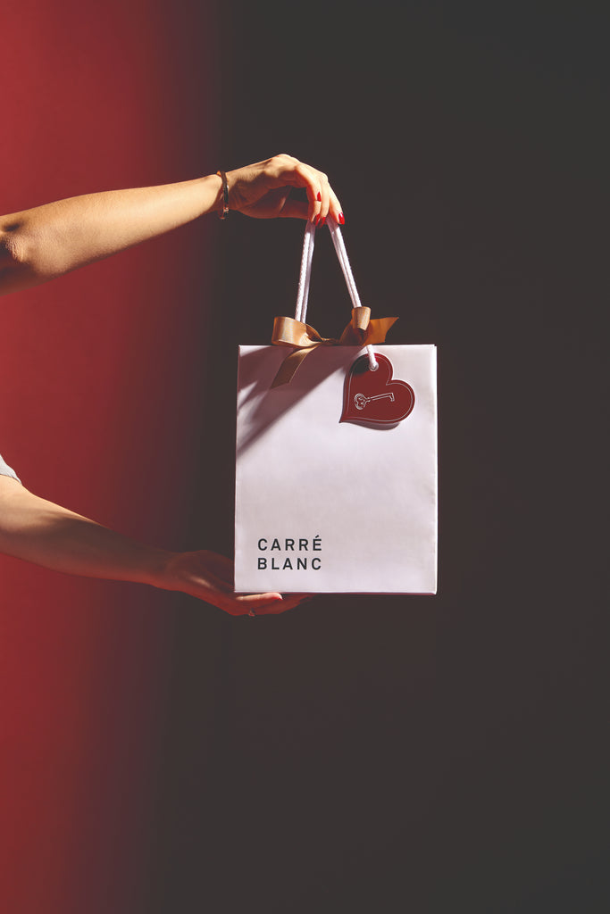 Person holding a white gift bag with 'Carré Blanc' branding against a dark background